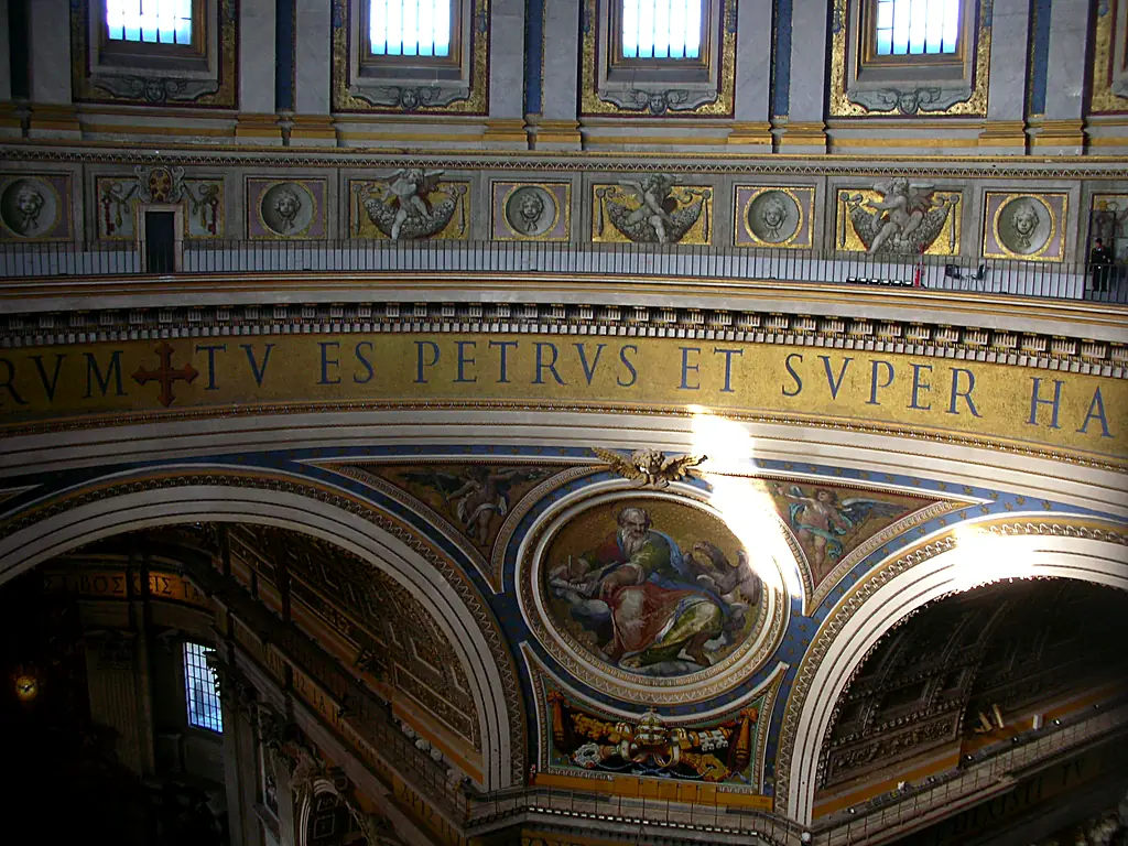 Ausschnitt des Kuppelfrieses: blaue Mosaikbuchstaben auf Goldgrund „…RVM ✠ TV ES PETRVS ET SVPER HA…“, darunter das Medaillon des Evangelisten Johannes mit dem Adler, darüber die Galerie und die Fenster des Tambours.